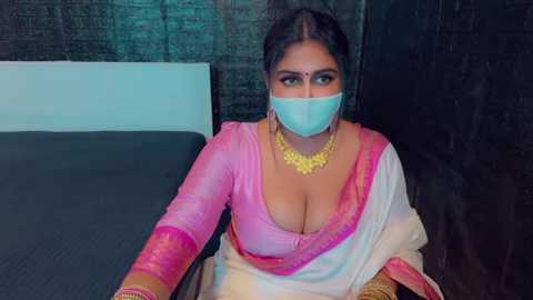 Media: Video of a South Asian woman in a pink saree with a plunging neckline, gold jewelry, and a blue surgical mask, seated on a dark bed with a light wall and textured black background.