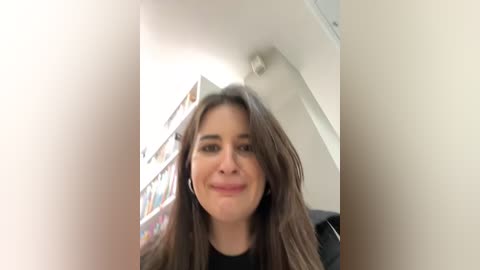 Media: Video of a smiling woman with long, wavy brown hair, wearing a black top, seated indoors near a bookshelf with colorful books.