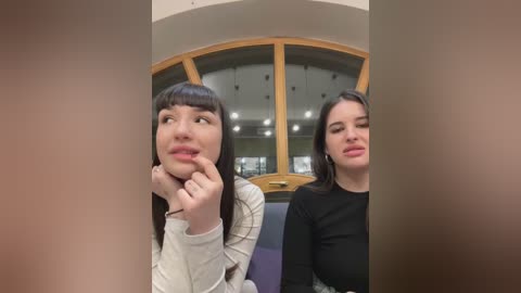 Media: Video of two young women with straight, dark hair, one in a beige sweater and the other in a black top, sitting in a modern, glass-enclosed room with wooden panels.