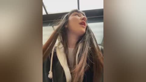 Media: A video of a young woman with long, straight, light brown hair, wearing a black jacket and a white scarf, with a blurred background suggesting an indoor setting.