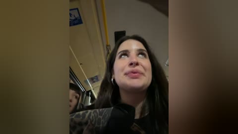 Media: Video of a young woman with long dark hair, wearing a black jacket, looking up with a thoughtful expression. Background features a yellow pipe and a wall with a blue sign.