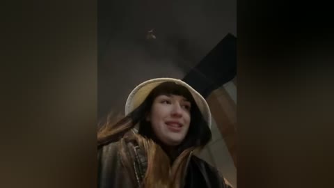 Media: Video of a smiling, light-skinned woman in a beige hat and brown coat, raising her gloved hand in the air, against a dark, blurry background.