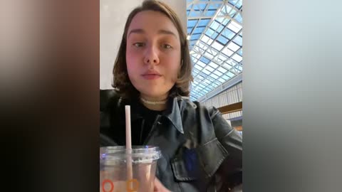 Media: Video of a young woman with shoulder-length brown hair, wearing a black jacket and holding a straw in a clear cup, in an indoor setting with a glass roof and metal beams.