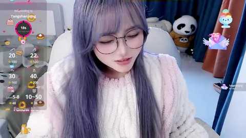 Media: Video of a young Asian woman with long, lavender hair, wearing glasses and a white sweater, sitting in a room with a panda plush toy and curtains.