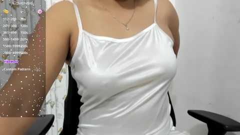 Media: Video of a light-skinned woman with a slender build, wearing a white spaghetti-strap tank top, sitting in a black office chair. Background shows a white wall and a floral-patterned curtain.