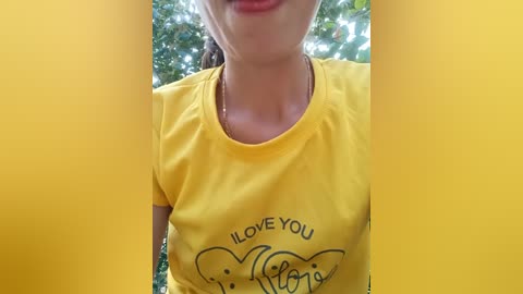 Media: Video of a light-skinned woman with a medium build, wearing a bright yellow t-shirt with \"I LOVE YOU\" in black text and a heart graphic. She is smiling slightly. Background features blurred green foliage.