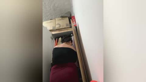 Media: Video of a woman with short brown hair, wearing a black top and red skirt, bending over to reach into a narrow, cluttered closet.