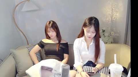 Media: Video of two young Asian women, one with long brown hair, in casual attire, sitting on a beige couch in a minimalist room with a curved floor lamp and flowers.