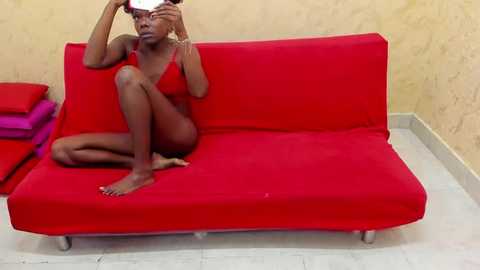 Media: Video of a dark-skinned woman with a slender build, wearing a red bra and panties, sitting on a red sofa in a plain, beige-walled room. She adjusts her hair with a mirror.