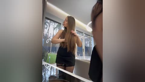 Media: A video of a young woman with long, straight brown hair, wearing a black sleeveless top and shorts, adjusting her hair in a modern, light-filled bathroom with large windows overlooking a lush garden.