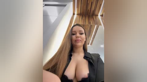 Media: Video of a confident, light-skinned woman with long brown hair, wearing a black open shirt, revealing large breasts, standing in a modern room with wooden beams and white walls.