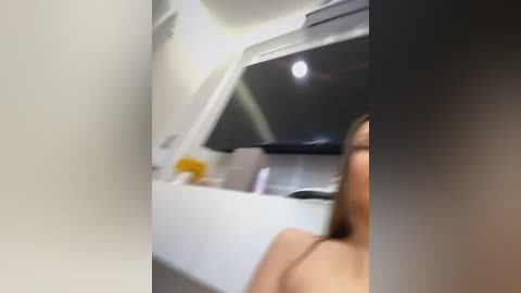 Media: Video of a blurred, partially visible woman with light skin and long brown hair, leaning over a white countertop in a modern kitchen, with white cabinets and a black appliance.