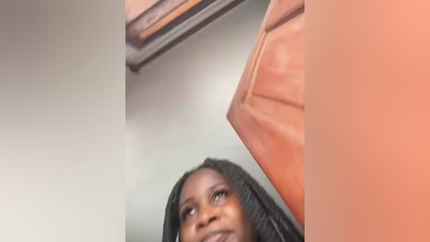 Media: Video of a young Black woman with long, braided hair looking upward, possibly in a bathroom with a beige ceiling and a wooden shelf.