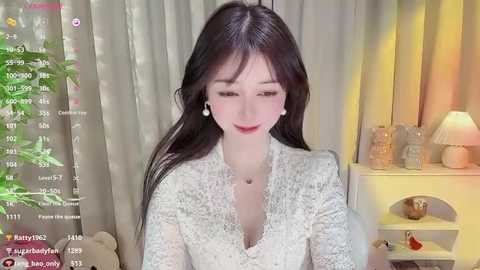 Media: Video of an East Asian woman with long black hair, fair skin, and a slim build, wearing a white lace dress, sitting in a room with a beige curtain, a lamp, and a plant.