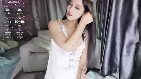 Media: Video of a young East Asian woman with long black hair, wearing a white lace camisole, adjusting her hair in a dimly lit living room with grey curtains and a white sofa.