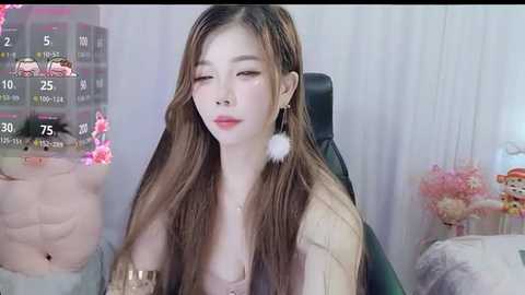 Media: A video of an East Asian woman with long brown hair, fair skin, and a slim physique, wearing a pink top and white fluffy earrings. She sits in a modern room with a gaming setup, floral decor, and a black gaming chair.