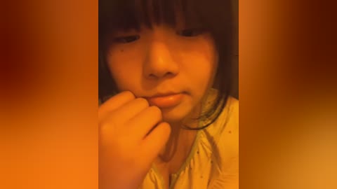 Media: Video of a young Asian girl with straight black hair and bangs, wearing a light-colored shirt. She is looking down, holding her hand to her mouth in a thoughtful pose. The background is blurred in warm colors.