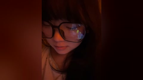 Media: A dimly lit video of a young Asian woman with straight, shoulder-length dark hair, wearing black-rimmed glasses, a white shirt, and a soft smile.