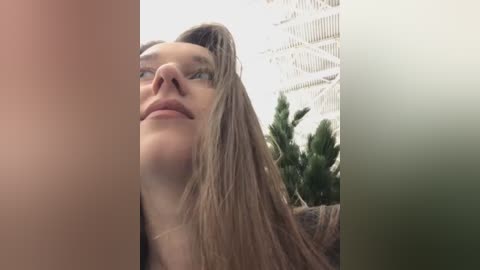 Media: Video of a young woman with long, straight blonde hair, fair skin, and blue eyes, captured from a low angle, with a blurred, modern indoor background featuring a white ceiling and greenery.