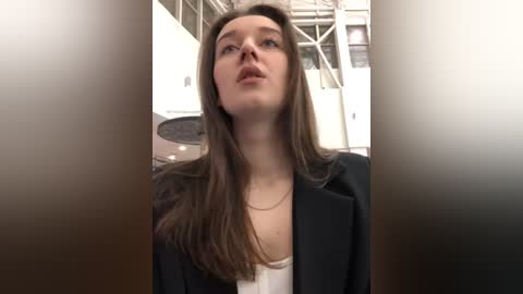 Media: Video of a young woman with long brown hair, wearing a black blazer, standing in a dimly lit, industrial space with metal structures and white walls in the background.