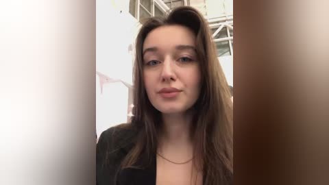 Media: Video of a young Caucasian woman with long brown hair, fair skin, and subtle makeup, wearing a black jacket, standing in a modern, white-walled hallway with metal railings.