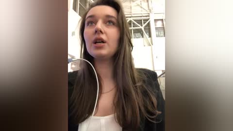 Media: A video of a young woman with straight, shoulder-length brown hair, wearing a white top and black jacket, listening to music with white earbuds. The background features a modern industrial setting with metal structures and glass windows.