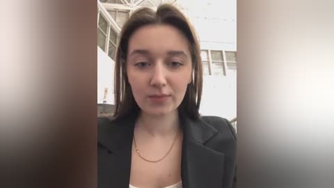 Media: A video of a young woman with fair skin, straight brown hair, and a neutral expression, wearing a black blazer over a white top. The background shows a white building with large windows and a metal structure.