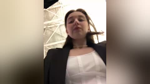 Media: Video of a young woman with straight, brown hair, wearing a white blouse and black blazer, standing indoors with a white geometric ceiling and a blurred person in the foreground.