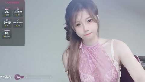 Media: Video of a young East Asian woman with long, straight brown hair, wearing a sheer, pink lace top, sitting indoors. The background is a blurred, light-colored room.