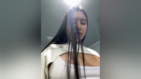 Media: Video of a woman with long, dark hair, wearing a white top, standing in a salon with dim lighting, hair being cut in a messy, uneven style.