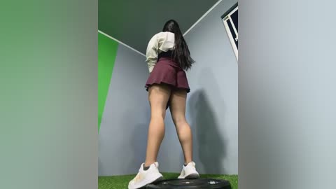 Media: Video of a young woman with long black hair, wearing a white blouse, maroon skirt, and white sneakers, standing on a green mat in a grey room with a window.