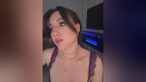 Media: Video of a fair-skinned woman with dark hair, wearing a lacy purple bra, pearls, and earrings, in a dimly-lit room with a TV and blue lights in the background.