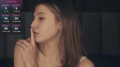 Media: A video of a young Caucasian woman with straight, medium-length brown hair, wearing a light-colored top, covering her mouth with her index finger, set against a dimly lit, blurred background.