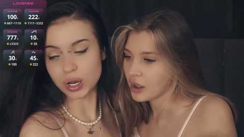 Media: Video of two young women with long hair, one wearing a pearl necklace, the other with a delicate necklace, both with closed eyes and relaxed expressions.