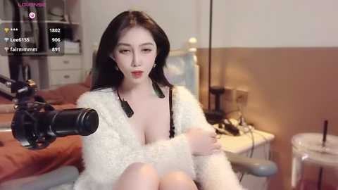 Media: Video of an Asian woman with long black hair, wearing a fluffy white robe, sitting on a bed with a black microphone in front of her, in a softly lit bedroom.