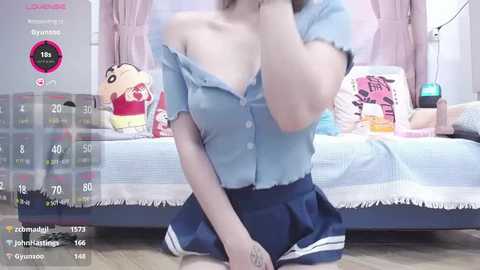 Media: Video of a slender, light-skinned woman with small breasts, wearing a blue off-shoulder top and navy blue skirt, sitting on a bed with pink curtains and plush toys.