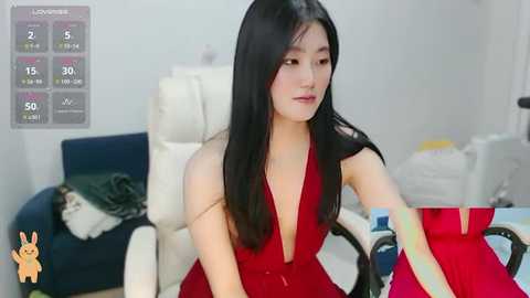 Media: A video of an Asian woman with long black hair and fair skin, wearing a red halterneck dress, sitting on a white chair in a cluttered room.