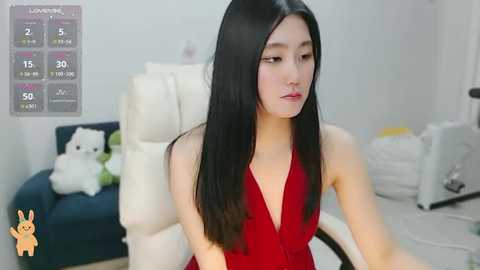 Media: Video of an Asian woman with long black hair in a red dress, sitting in a white chair. Background shows a calendar, stuffed animal, and cluttered room.