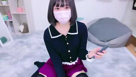 Media: Video of an Asian woman with a short black bob haircut, wearing a navy blue cardigan, pink mask, and purple skirt, kneeling on a gray carpeted floor, holding a remote.