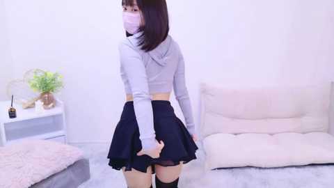 Media: Video of an Asian woman with a light complexion, wearing a light grey crop top, black skirt, and thigh-high socks, adjusting her skirt, standing in a minimalist, white room with a beige chair and a white table.