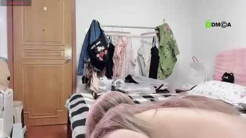 Media: A video of a messy bedroom with a person's legs in striped socks in the foreground, a bed with a pink blanket, and hanging clothes on a rack in the background.