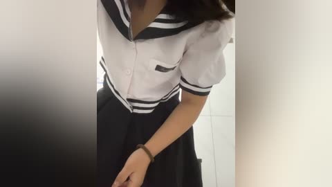 Media: Video of a woman in a black-and-white sailor school uniform, with a white shirt and black pleated skirt, standing in a tiled bathroom.