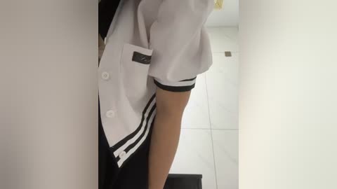 Media: Video of a person in a white, short-sleeved shirt with black trim, black skirt, and black shoes, standing in a white-tiled corridor. The image focuses on the legs and torso.