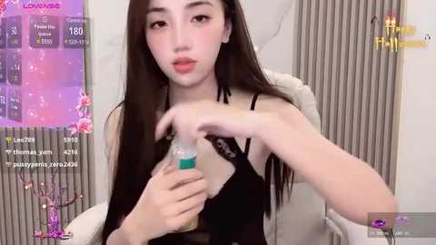 Media: Video of a young Asian woman with long black hair, fair skin, and a slender build, wearing a black top, holding a blue and white drink bottle. The background features a modern room with muted tones and a pink and purple digital overlay.