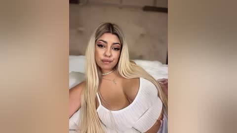 Media: Video of a blonde woman with long hair, large breasts, wearing a white, low-cut top, lying on a bed with white sheets and a beige headboard.