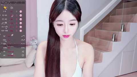 Media: A video of an East Asian woman with long, straight black hair in a white halter top, sitting on a white staircase with a plush toy, in a modern, minimalistic room.