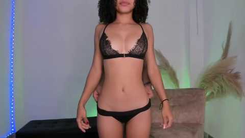 Media: Video of a slender Black woman with curly hair wearing black lace lingerie, standing in front of a beige couch with pampas grass and blue LED lights in the background.