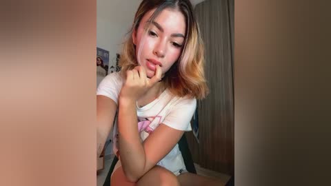 Media: Video of an Asian woman with shoulder-length, wavy, light brown hair, wearing a white T-shirt and shorts, sitting in a room with beige walls and wooden furniture, making a pouty face.