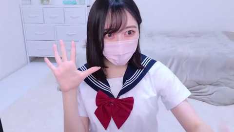 Media: Video of an East Asian woman with straight black hair, wearing a schoolgirl uniform with a red bow, white mask, and waving in a bright, minimalistic bedroom with white furniture.