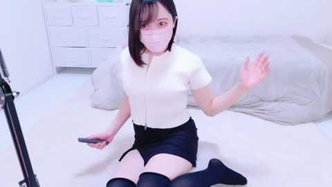 Media: Video of an Asian woman with straight black hair, fair skin, and a slim build, wearing a white blouse, black shorts, and knee-high black socks, kneeling on a white floor in a minimalist room with a white dresser and bed.